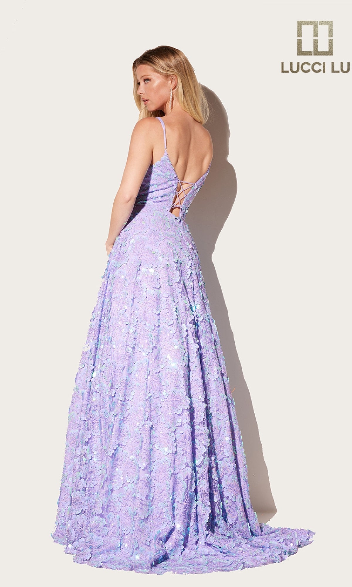 A woman in the Lucci Lu 1325 long prom dress with pockets, featuring a lavender color, spaghetti straps, embellished details, and a lace-up back stands against a plain light background with the Lucci Lu logo.
